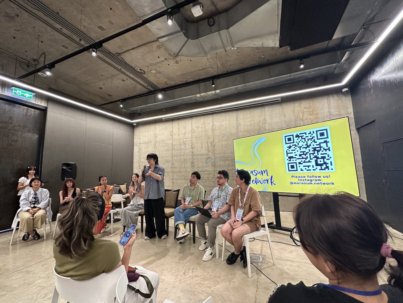 A row of people speaking in a room in front of a projector screen. A row of people speaking in a room in front of a projector screen.