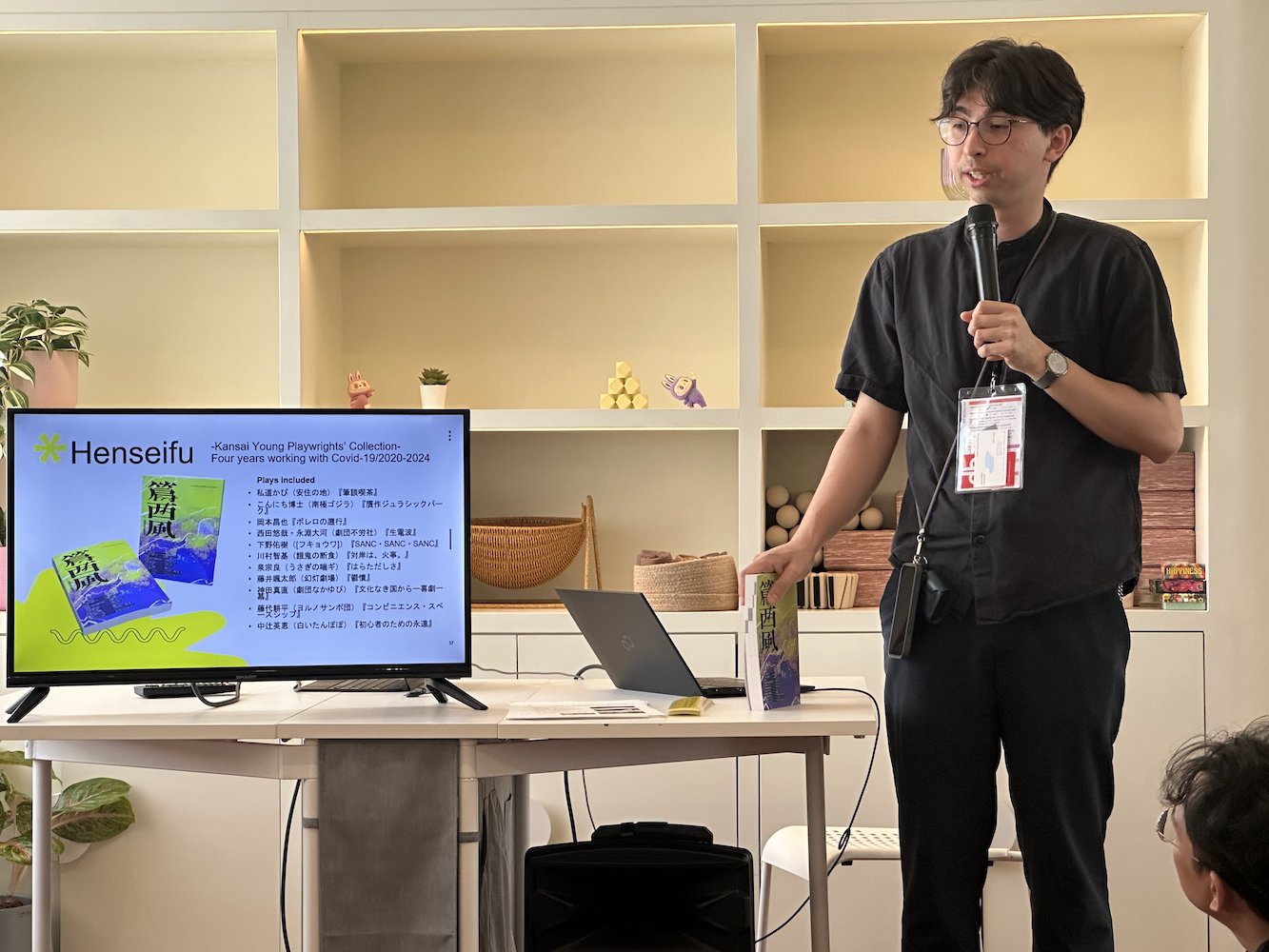 A male-presenting person speaking into a microphone. Beside him, on a table, is a TV screen. A male-presenting person speaking into a microphone. Beside him, on a table, is a TV screen.