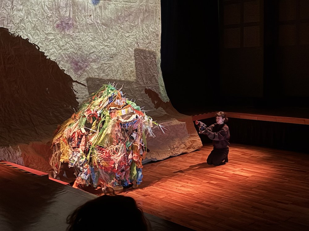 A large moving puppet made out of plastic trash, bent in front of a puppeteer wearing black. A large moving puppet made out of plastic trash, bent in front of a puppeteer wearing black.