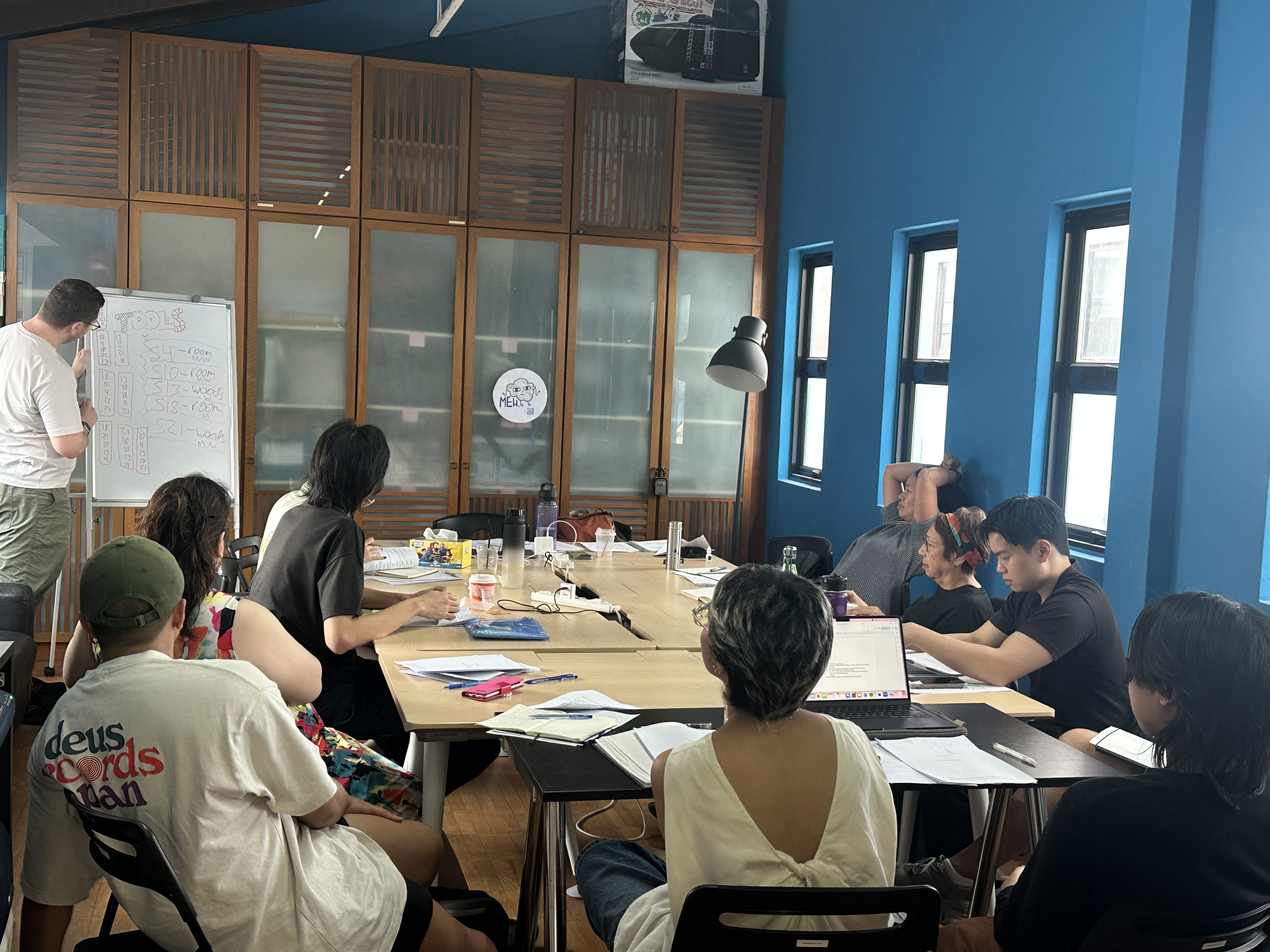 A photograph of the workshop with Sean by the whiteboard where there a few number sequences written and participants seated around the tables looking in his direction.