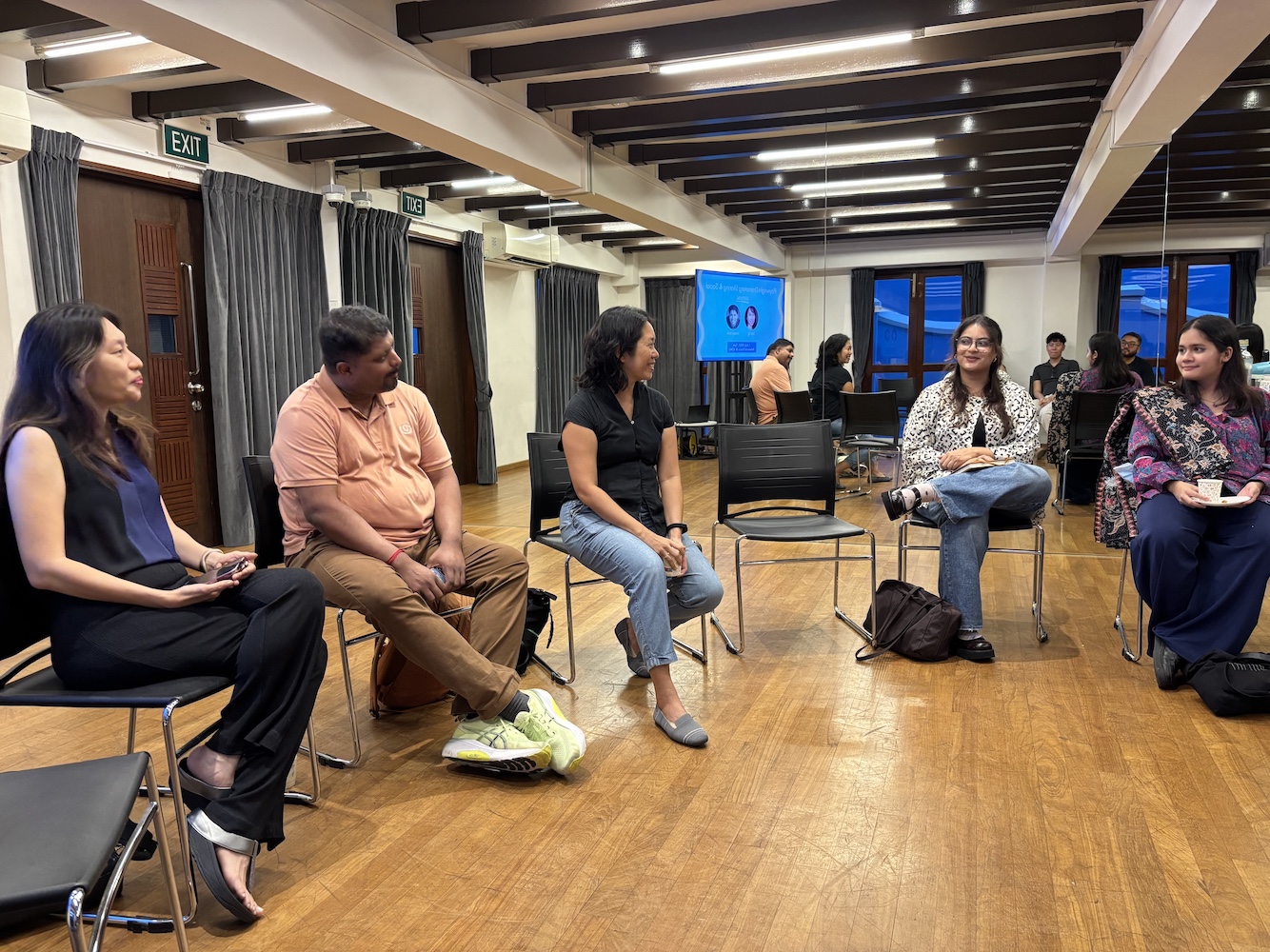 Five people seated on chairs, in front of a large mirror, speaking to each other.