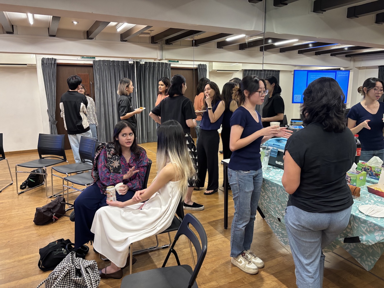Several people standing and sitting in a small room, talking to one another in small groups.