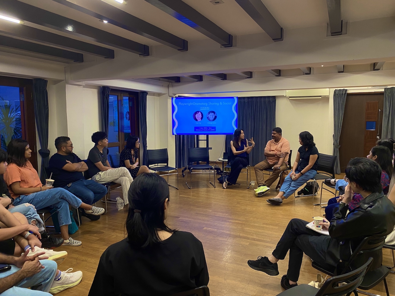 A group of people seated in a circle in a small room. In the corner is a TV monitor.