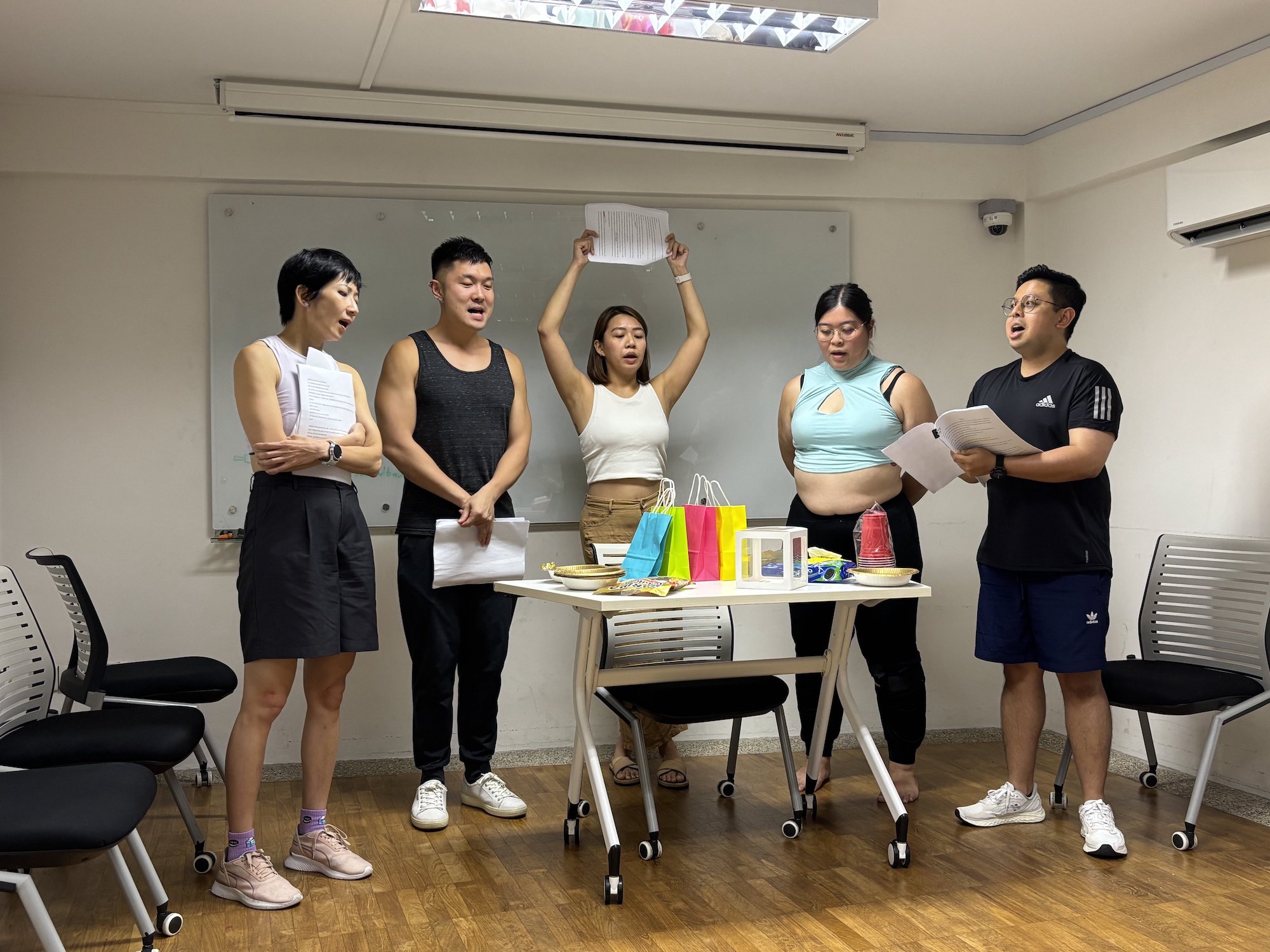 Five persons standing around a small table laden with objects, singing.