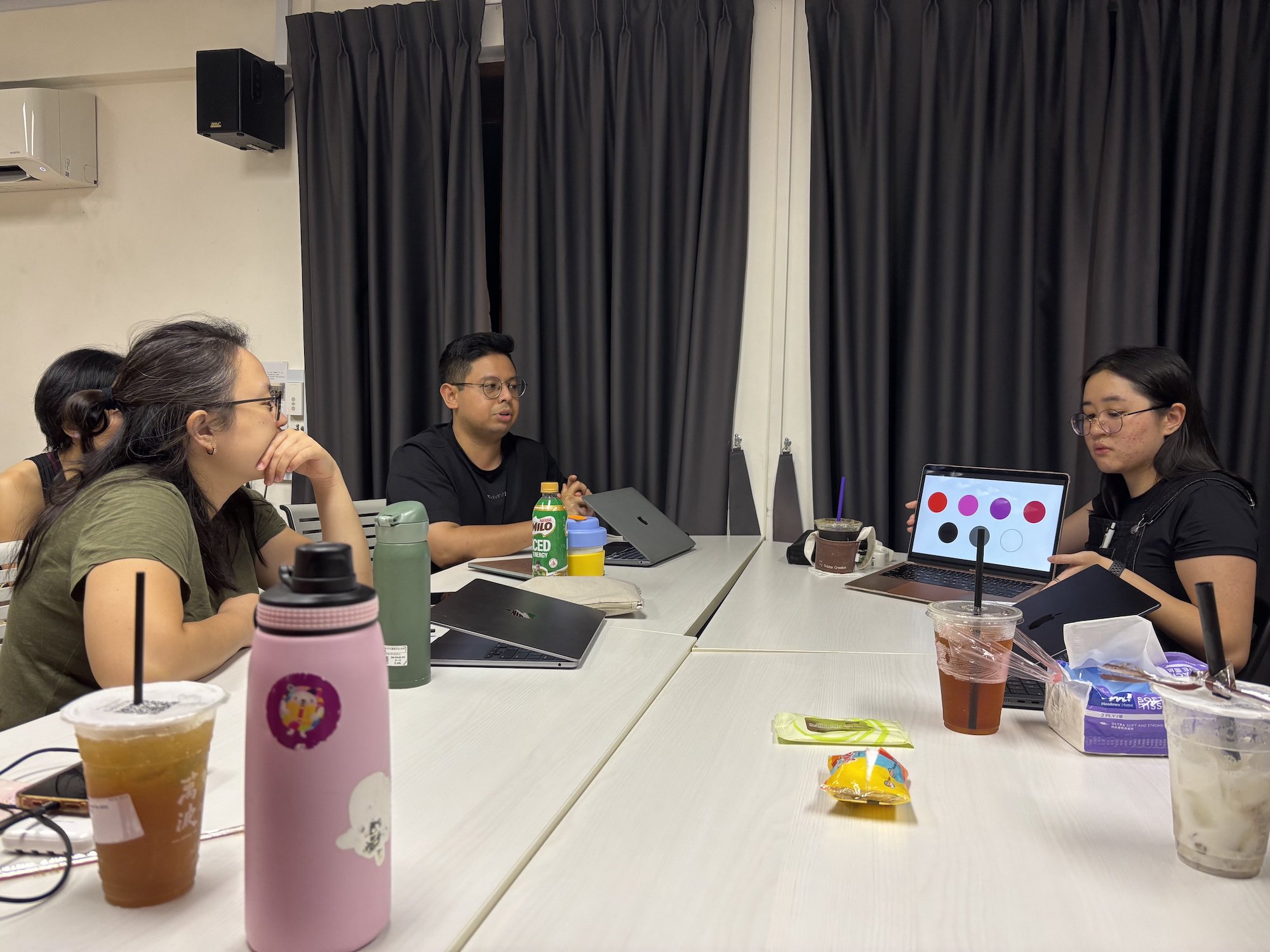 Three persons seated around white tables, talking over a laptop.