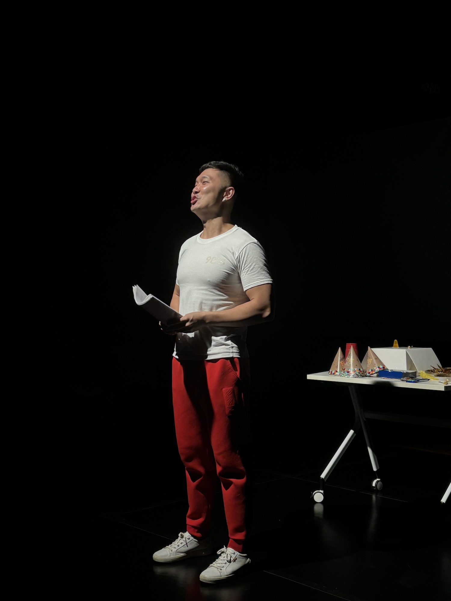 A male-presenting person wearing a white shirt and red pants, holding a script and speaking in the dark.