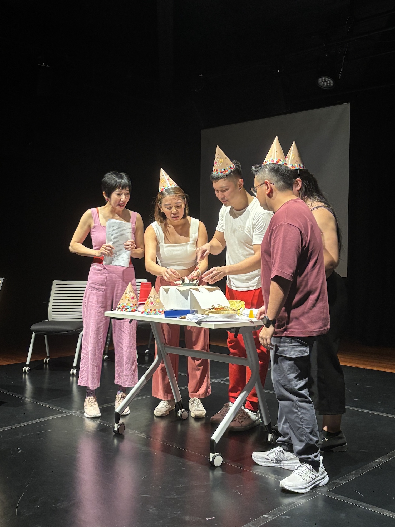 Five persons in party hats standing around a small table, cutting a very small cake together.