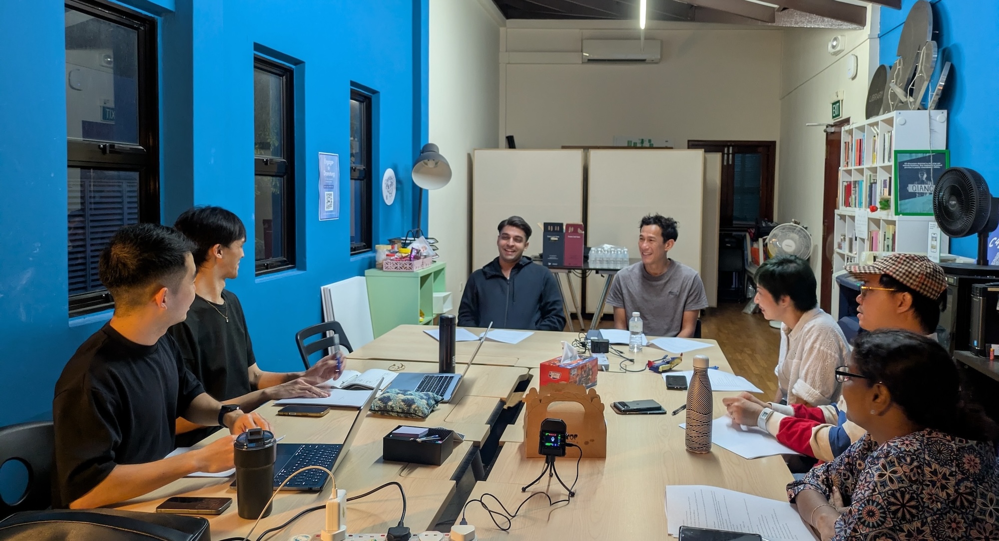 Seven people seated around a cluttered table, talking to each other. Seven people seated around a cluttered table, talking to each other.