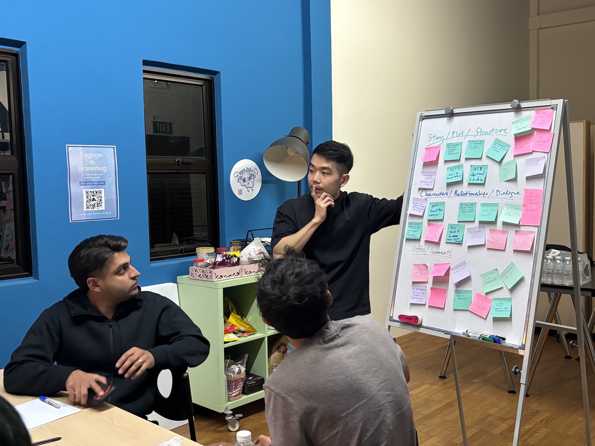 A male-presenting person stands beside a whiteboard filled with post-its, looking thoughtful. In front of him, two other people stare at the whiteboard. A male-presenting person stands beside a whiteboard filled with post-its, looking thoughtful. In front of him, two other people stare at the whiteboard.