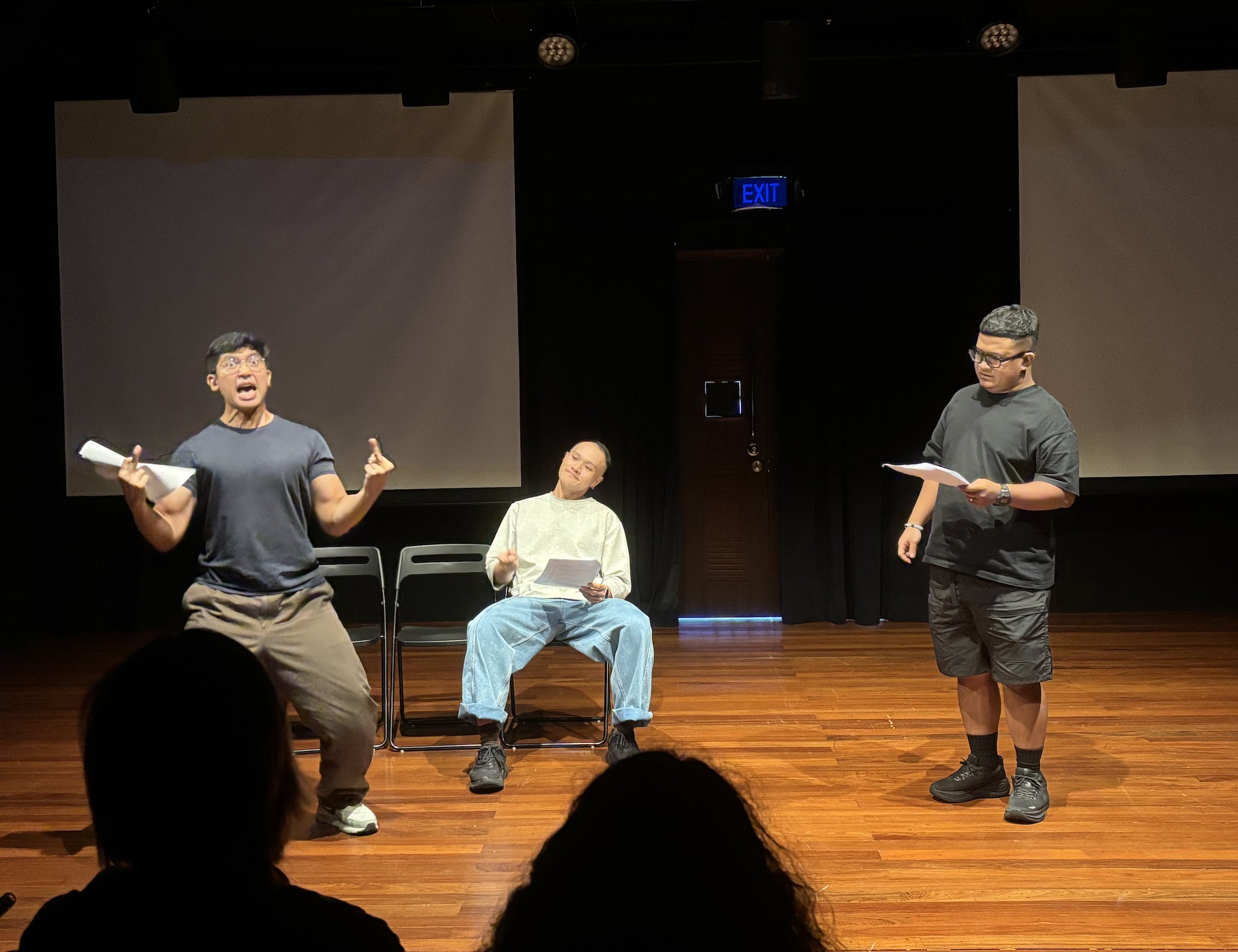 Three male-presenting persons, one flipping the bird, one seated and smiling, and one standing and observing quietly. Three male-presenting persons, one flipping the bird, one seated and smiling, and one standing and observing quietly.