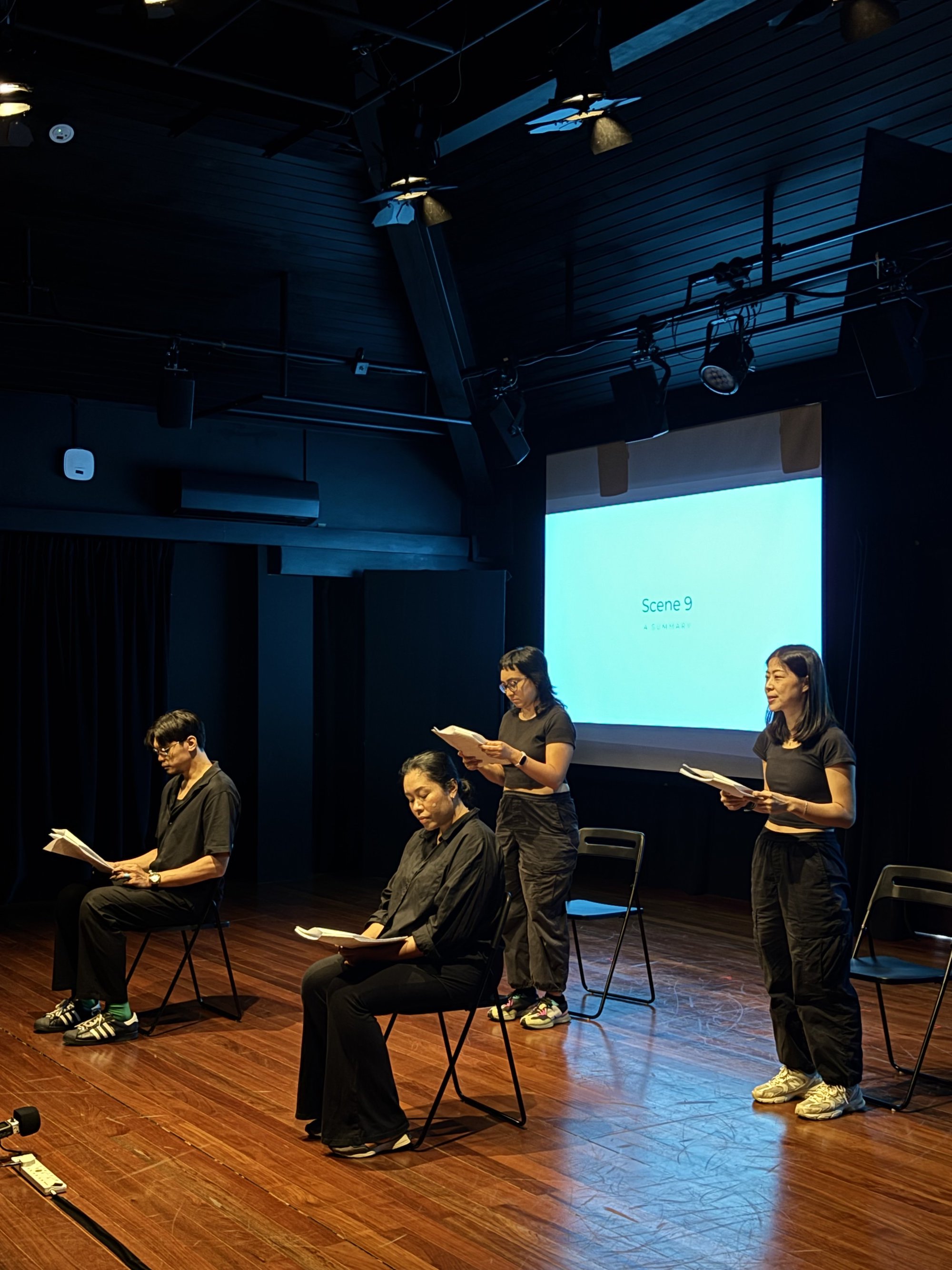Four persons seated and standing under a spotlight, reading from scripts.