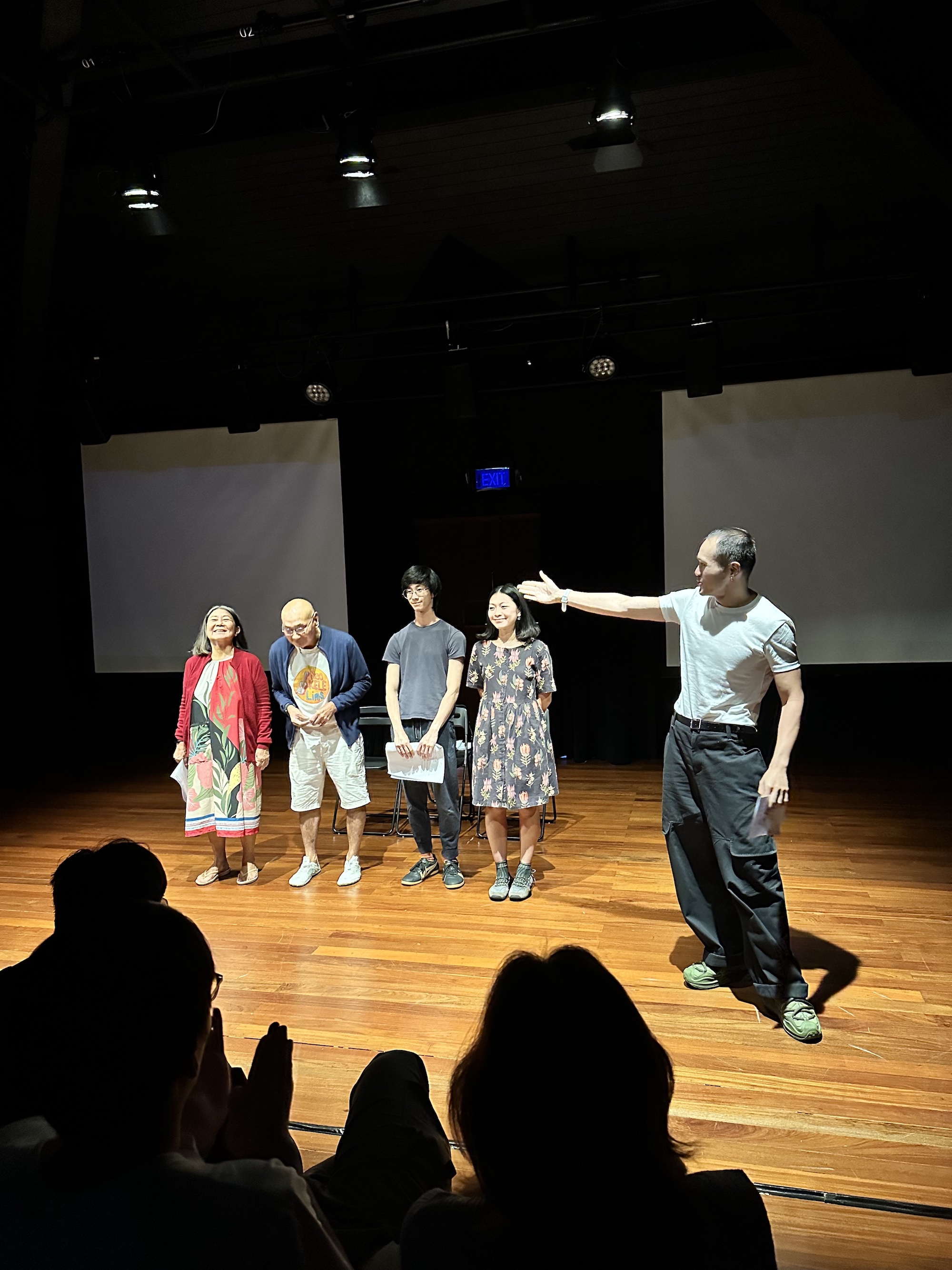 Four persons standing and smiling. Another person to the left gestures widely at them.