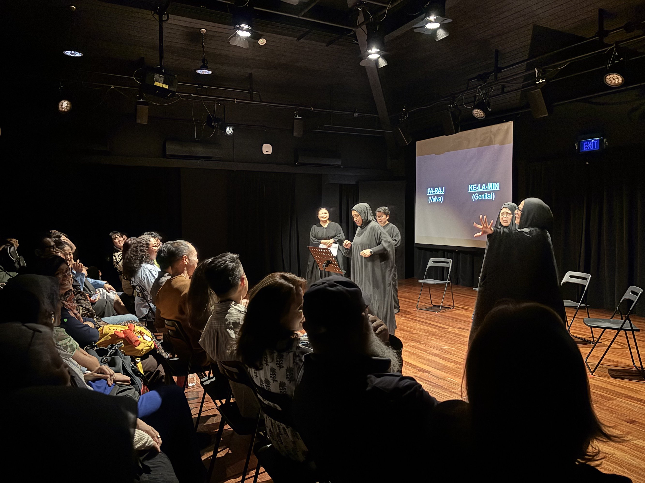 Five female-presenting persons in long black abayas, gesturing to an audience in a black box theatre.