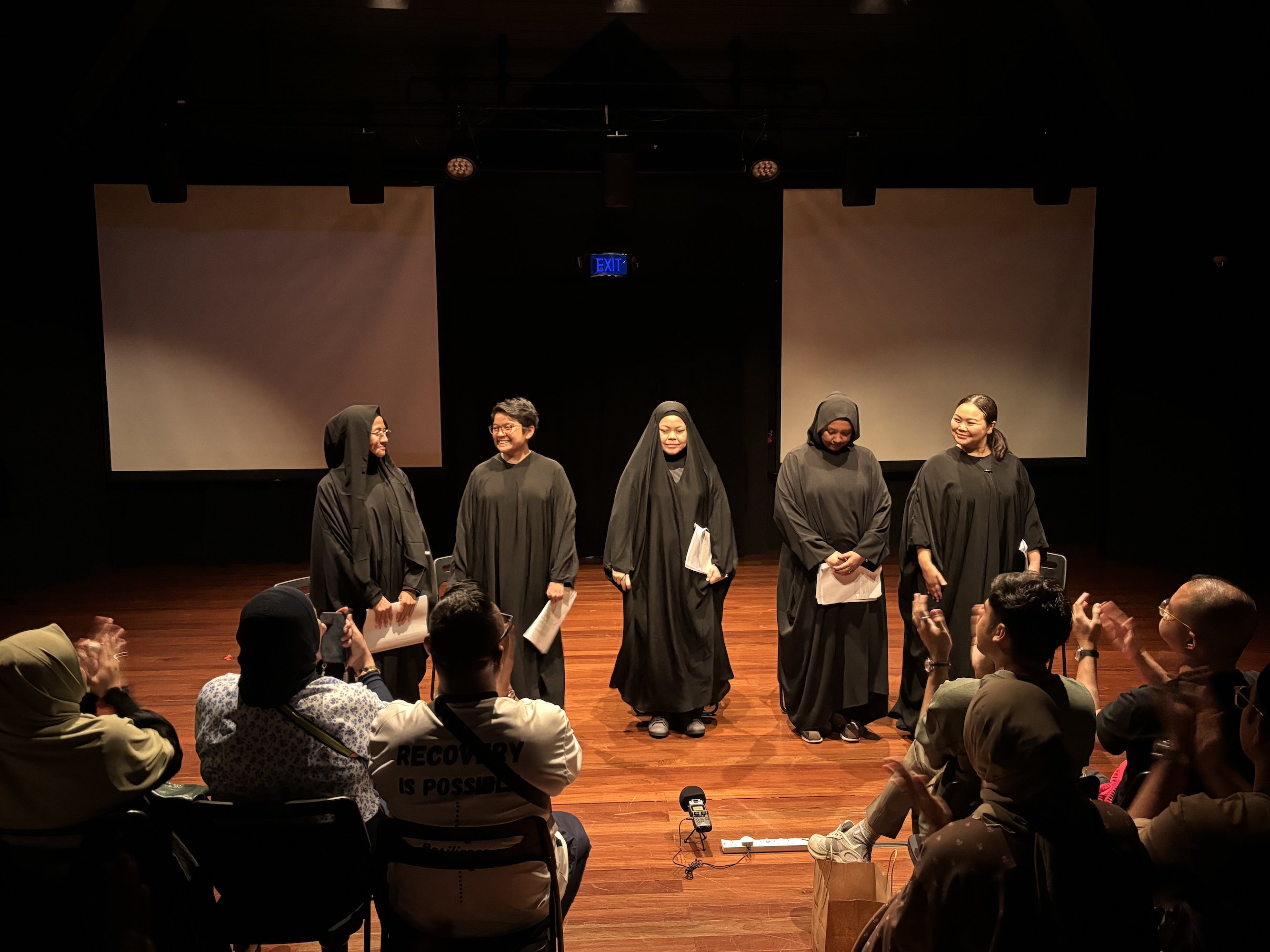 Five female-presenting persons in long black abayas standing in a row in front of an audience, smiling.