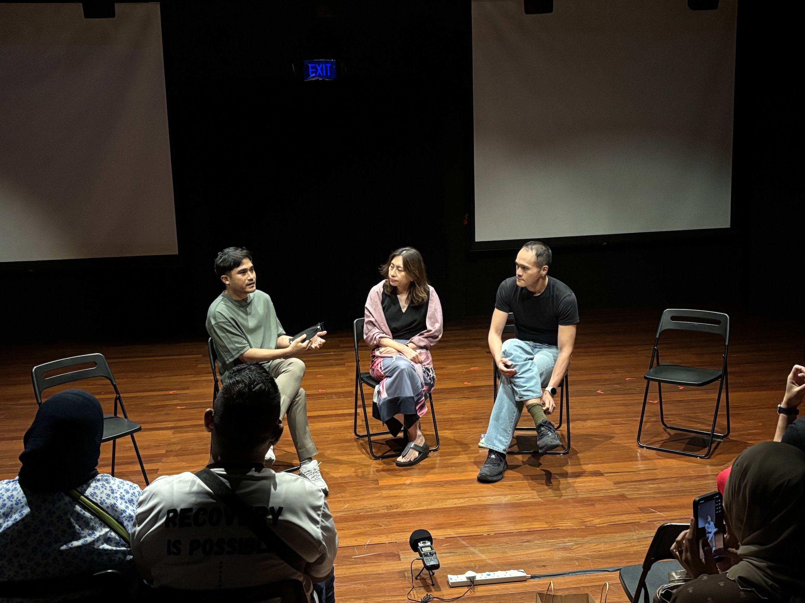 Three persons seated in chairs, spotlit, in front of an audience.