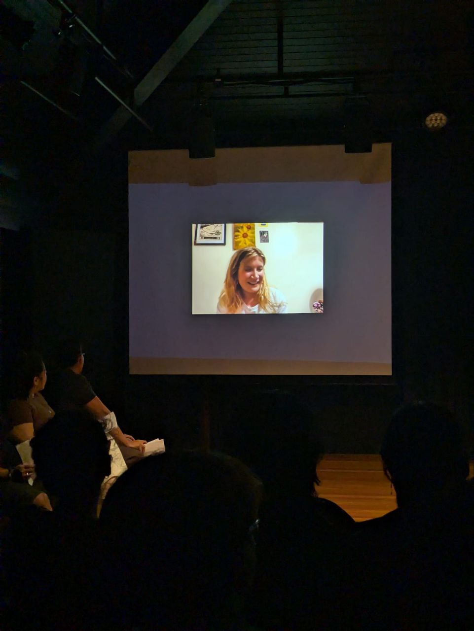 A smiling female-presenting person in a video projected on a screen. A smiling female-presenting person in a video projected on a screen.