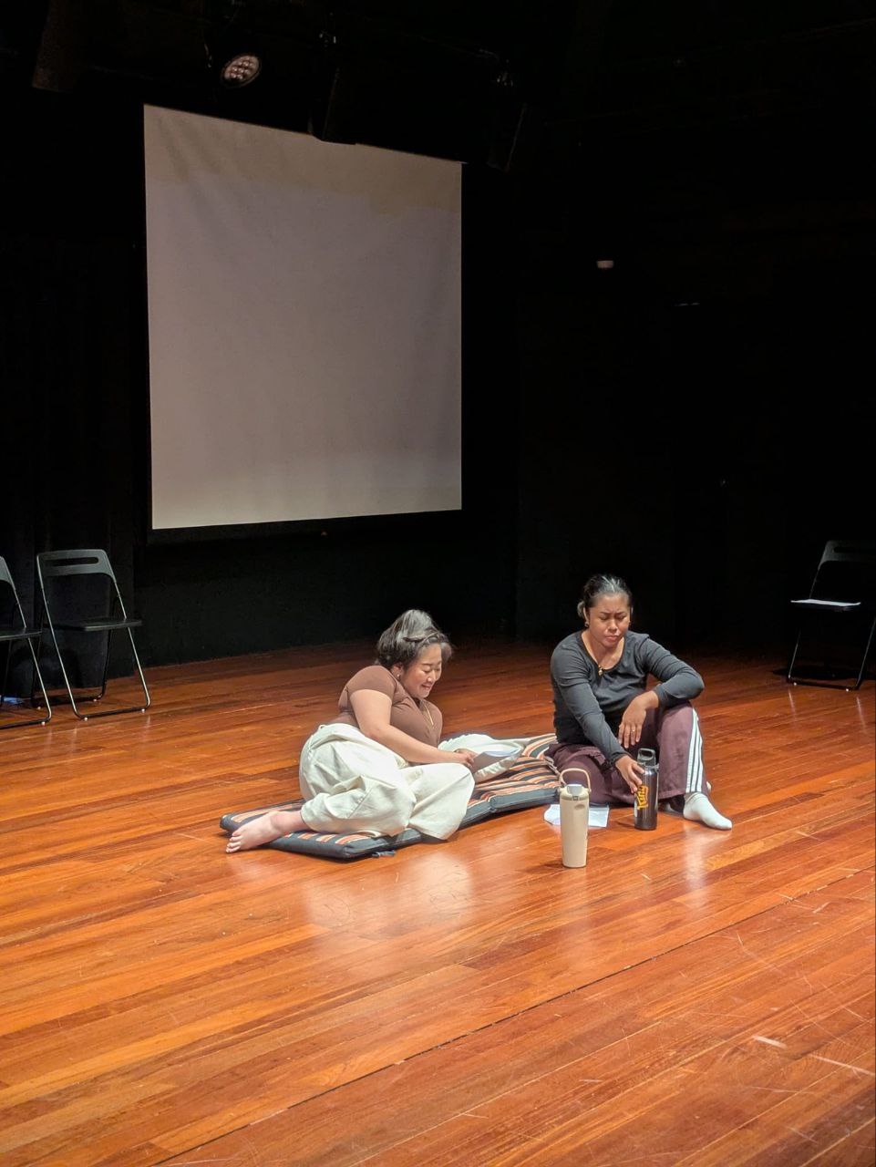 Two female-presenting persons seated on the wooden floor of a black box. Two female-presenting persons seated on the wooden floor of a black box.