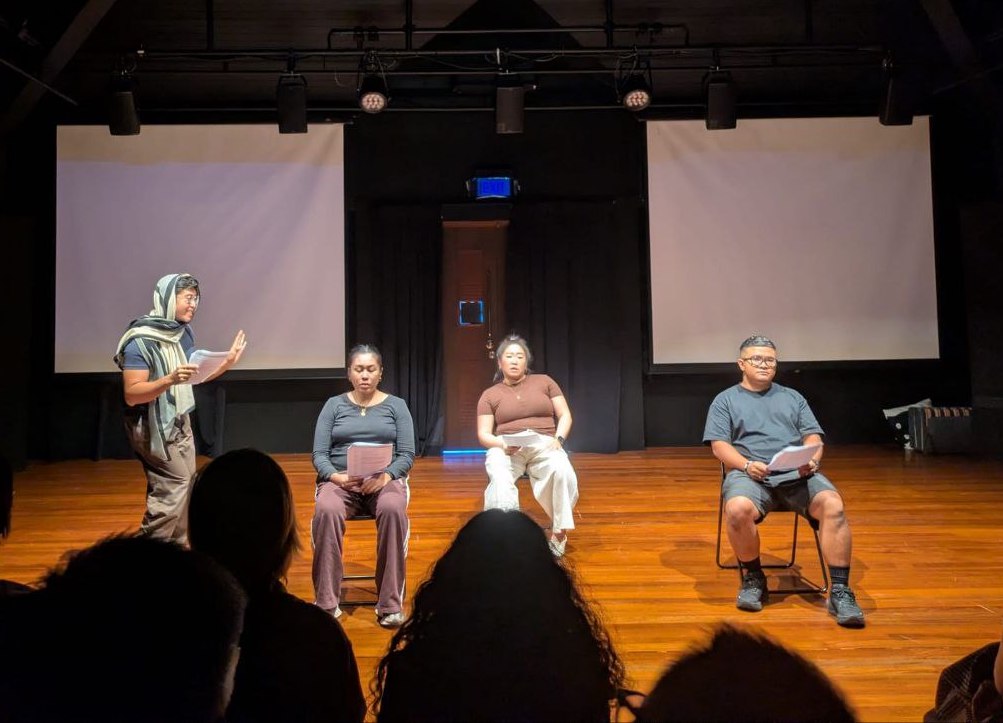 Three people seated in chairs, staring blankly. Another person in a headscarf stands and watches them. Three people seated in chairs, staring blankly. Another person in a headscarf stands and watches them.
