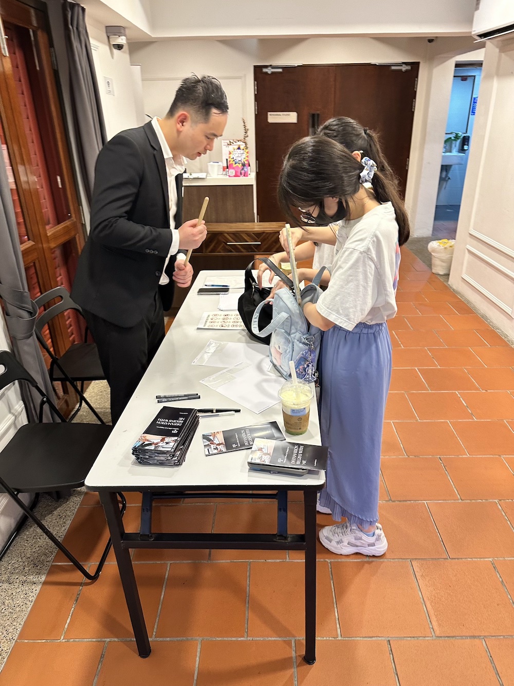 A male-presenting person in a suit checking two persons' bags at a table.