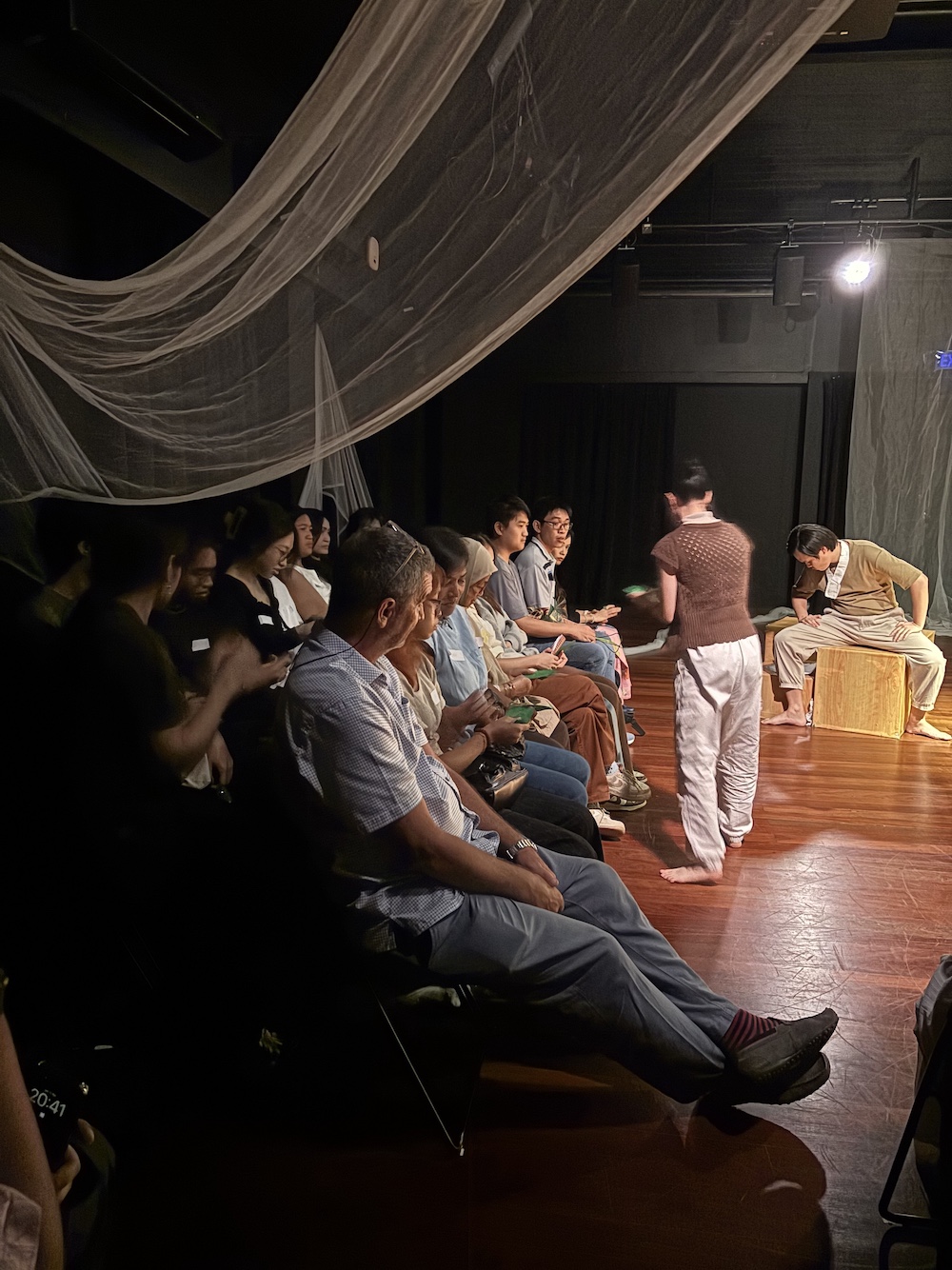 Two rows of audiences seated in a black box. Draped above them are thin translucent sheets of fabric.
