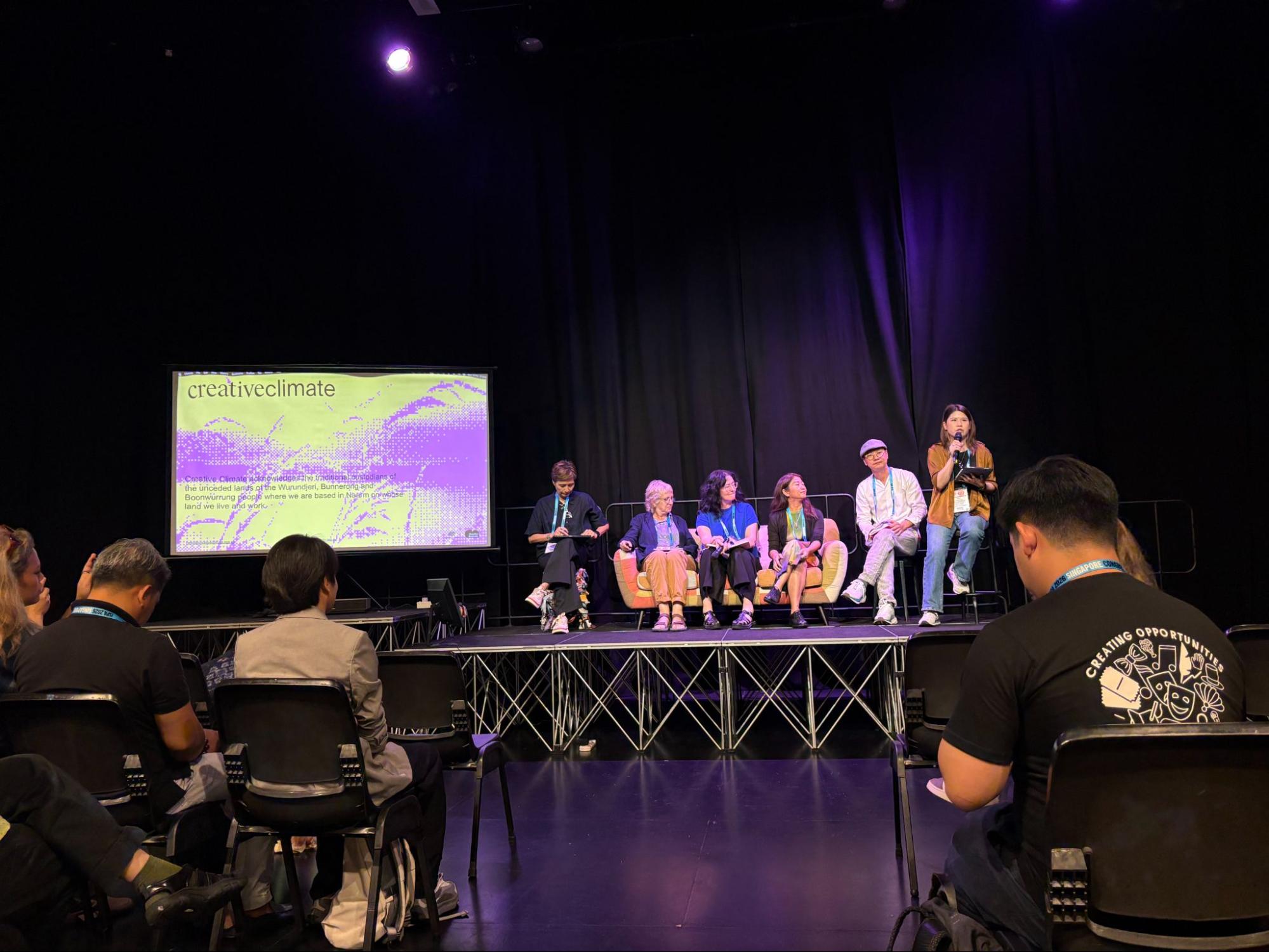 Six people perched on a couch on a stage.