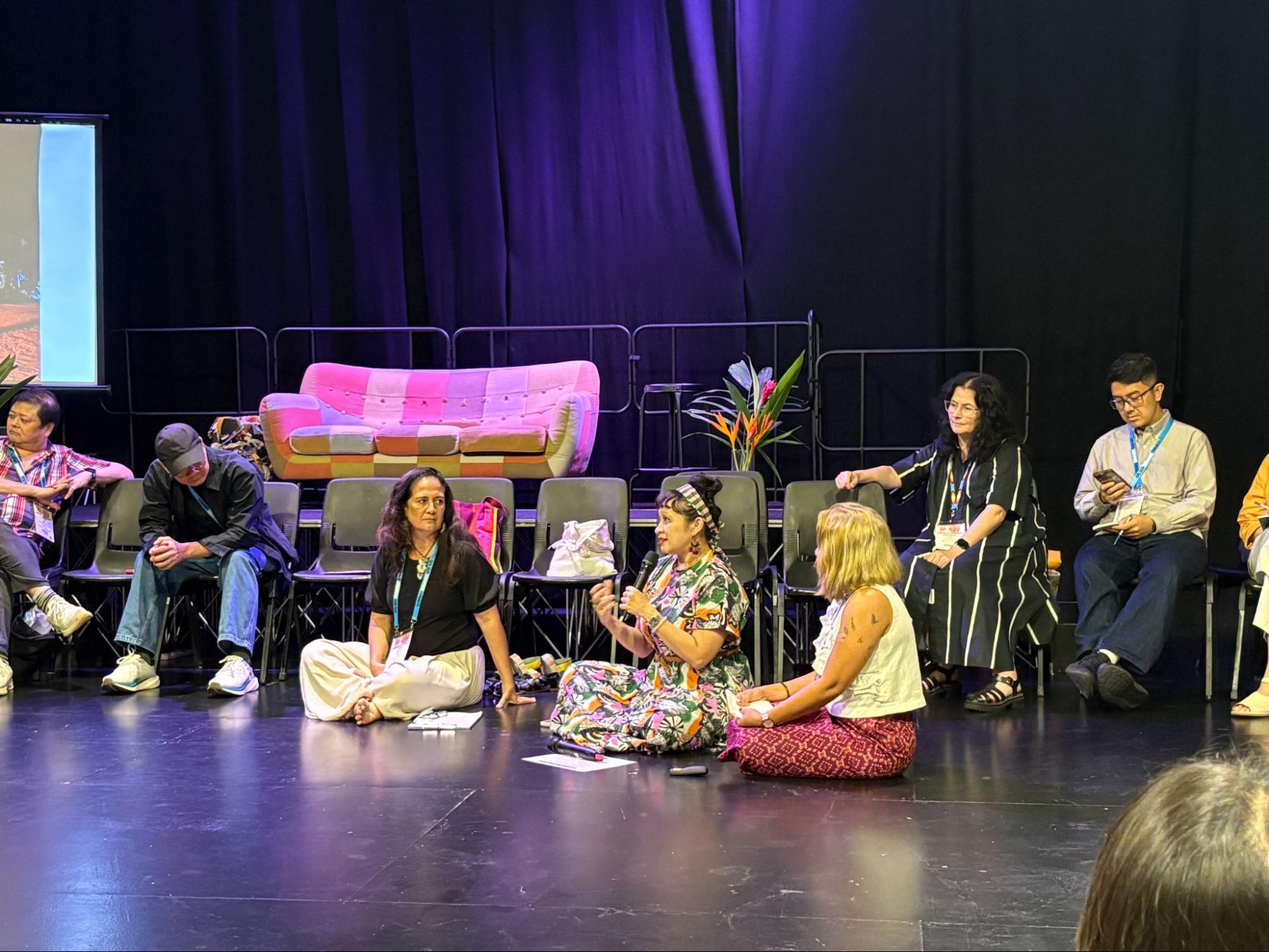 A few female-presenting persons seated on the floor. One speaks into a microphone.