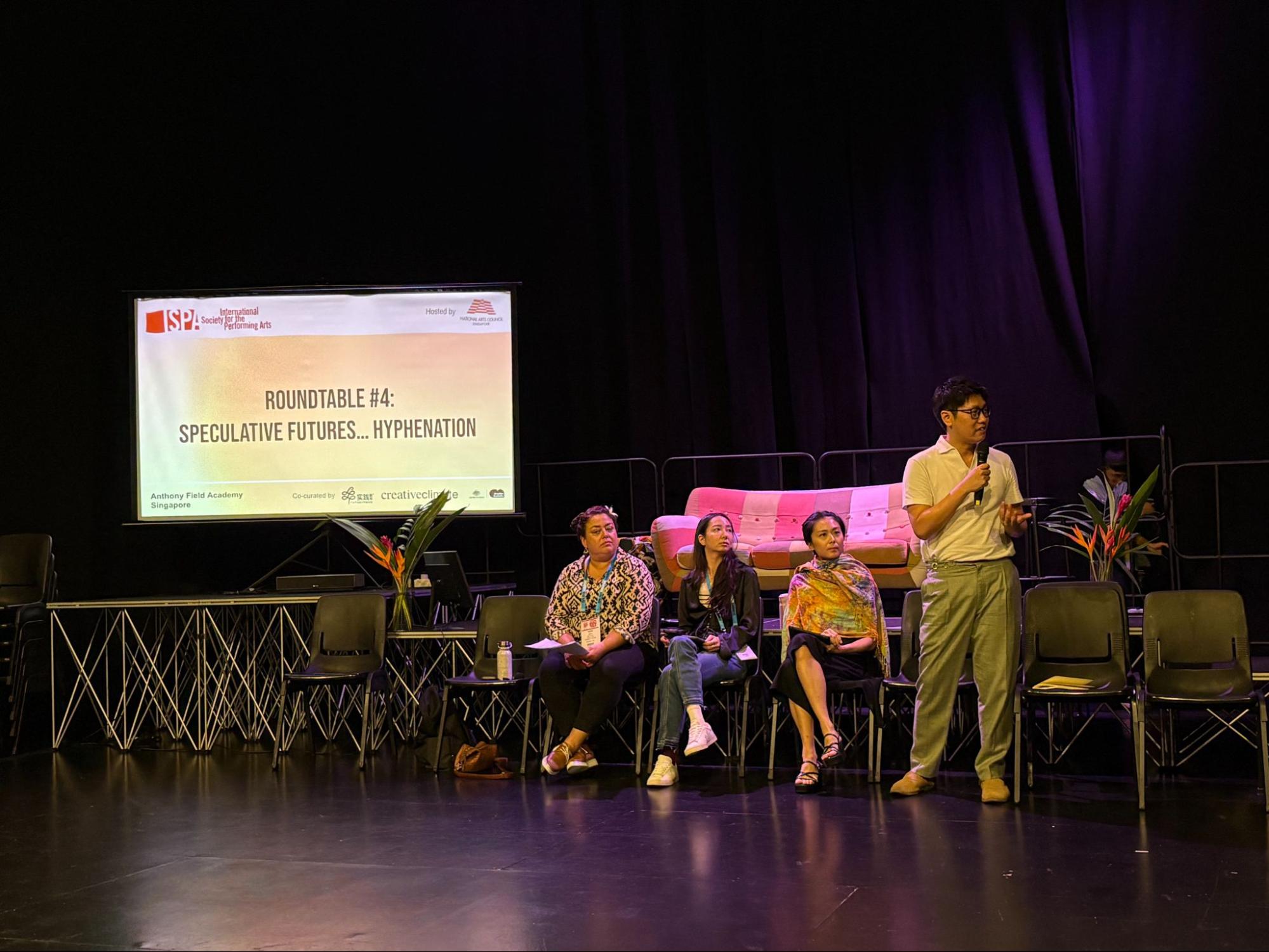 Three persons seated on chairs. To their left stands a male-presenting person speaking into a microphone.