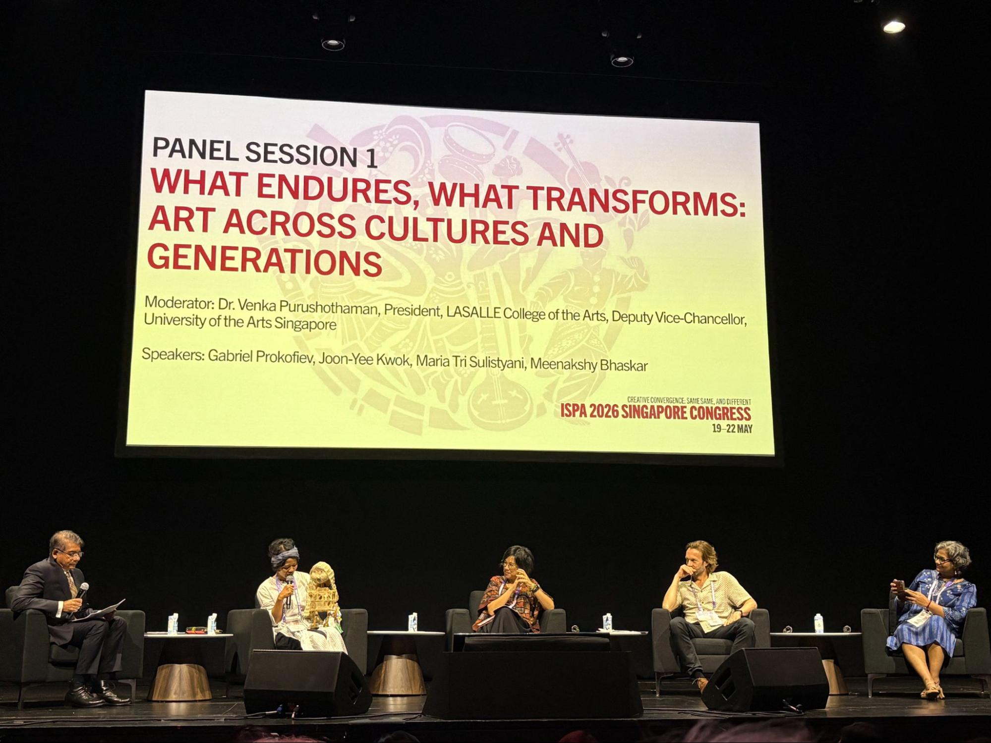 Five persons seated on a stage, under a large screen.