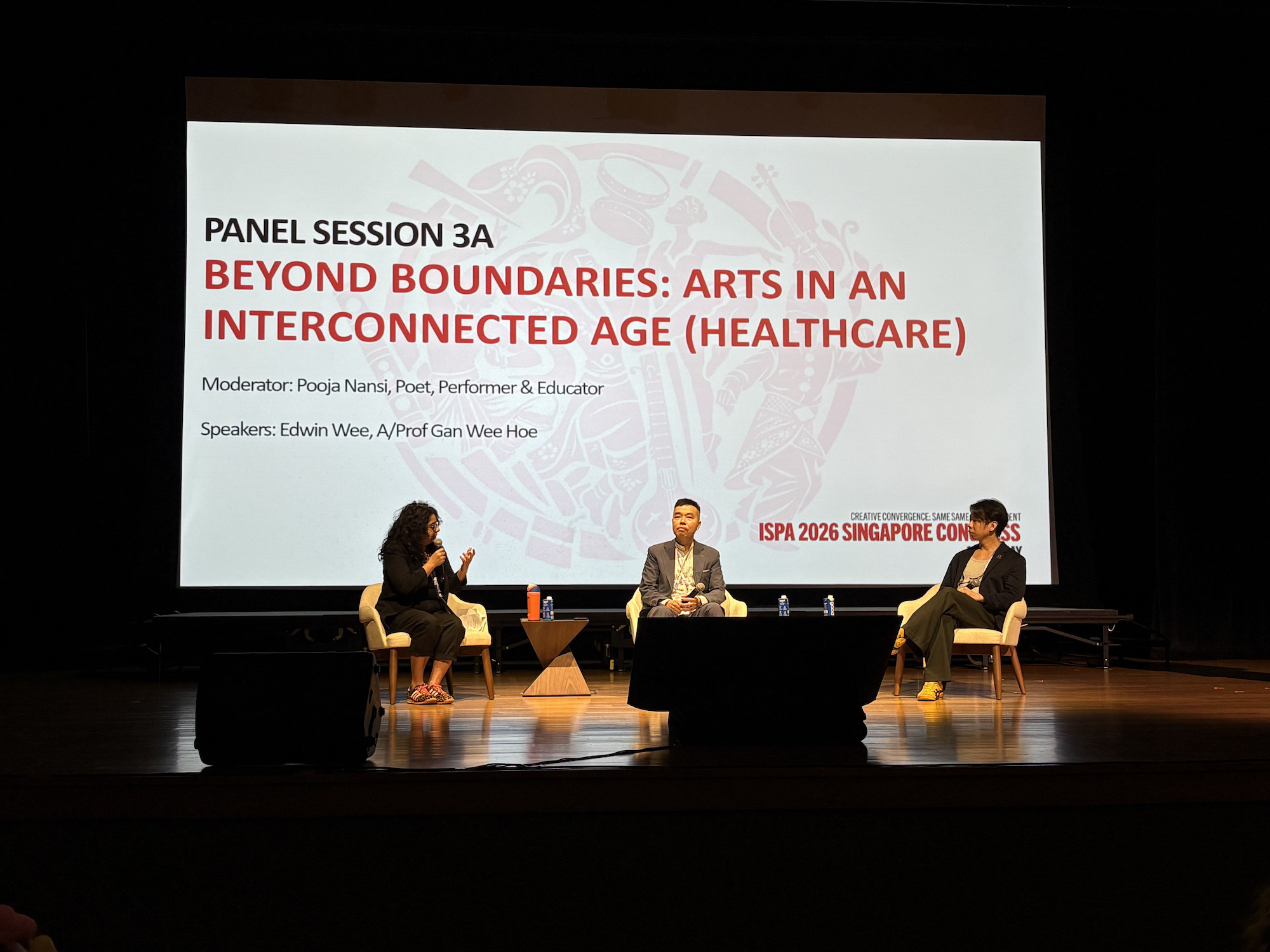 Three persons seated on a stage. Behind them is a large projector screen.
