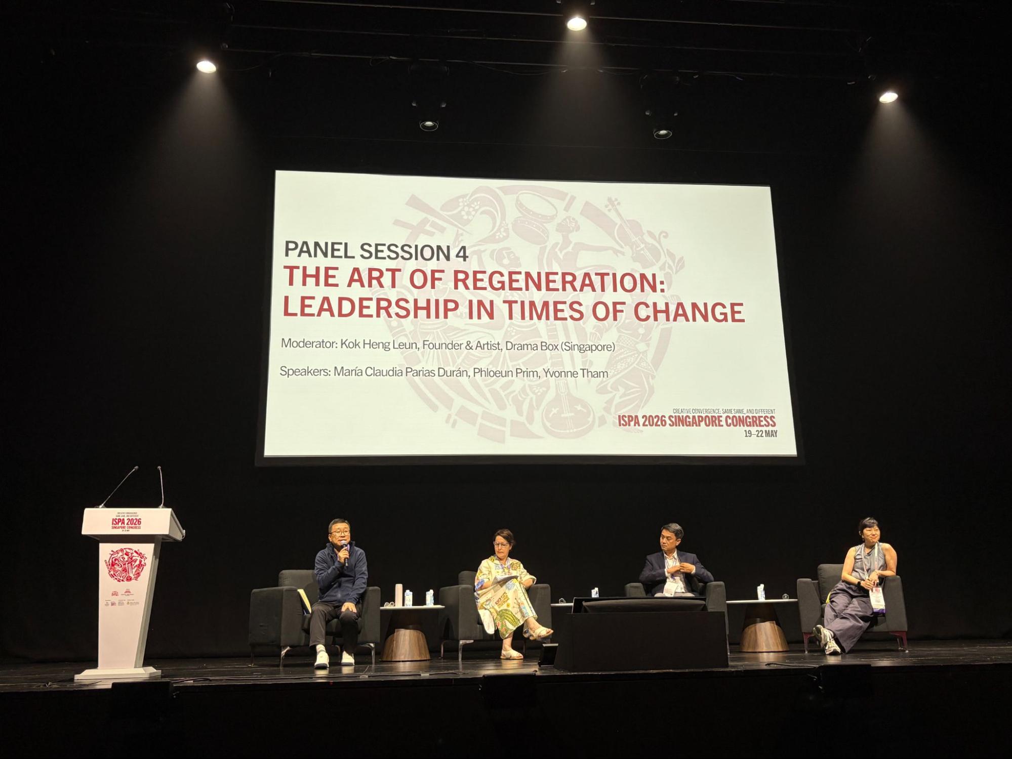 Four persons seated on a stage, under a large screen.