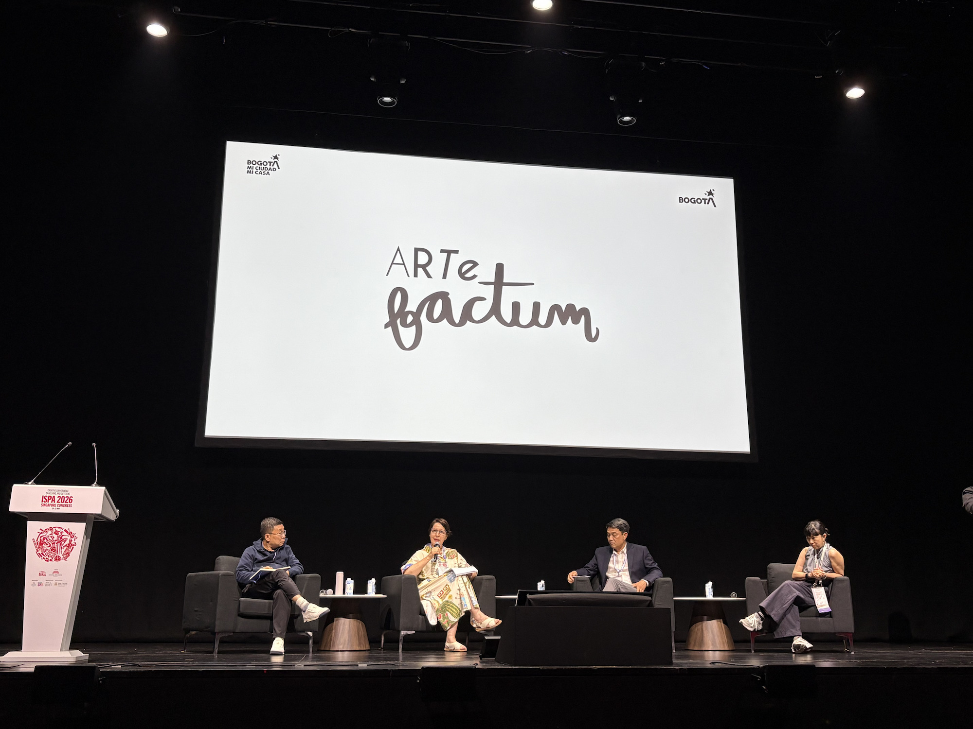 Four persons seated on a stage under a large screen, which reads 'Artefactum'.