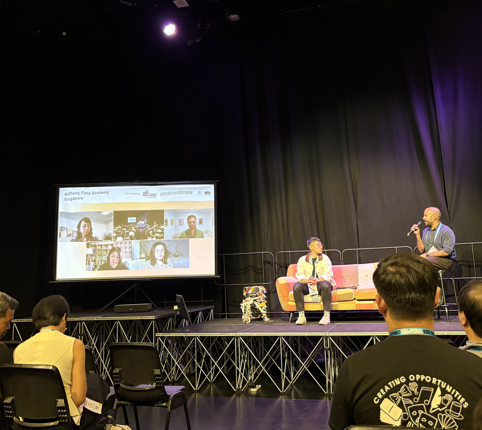 Two people seated on a couch on a stage. To the left is a monitor featuring five people on Zoom.