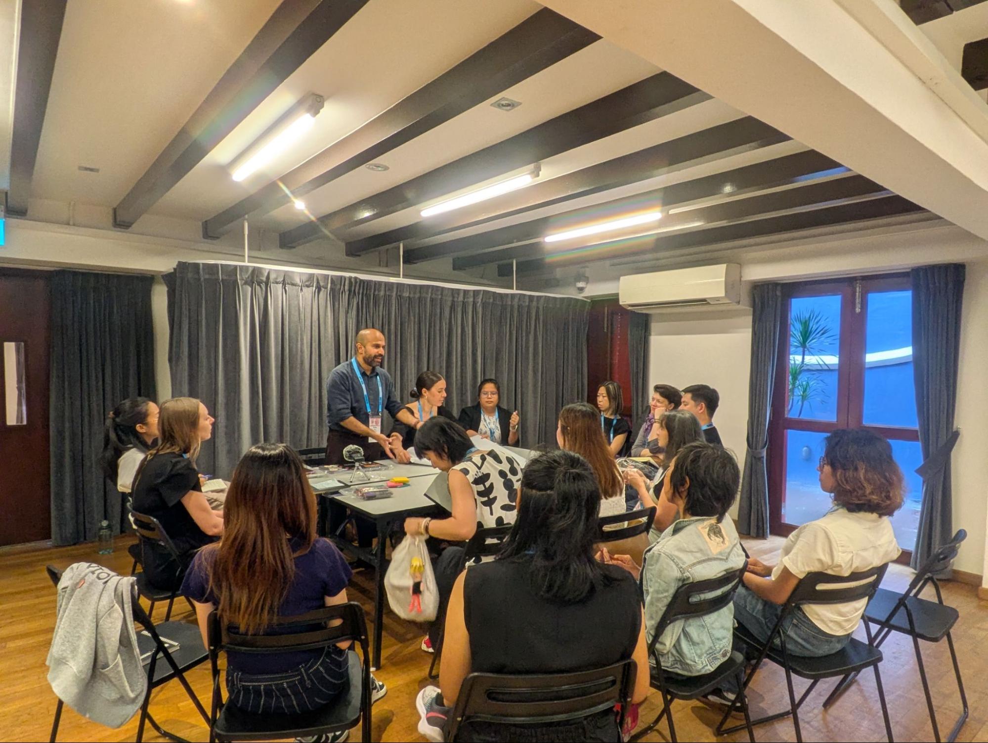 A group of people seated around a table. A male-presenting person stands and speaks to them.