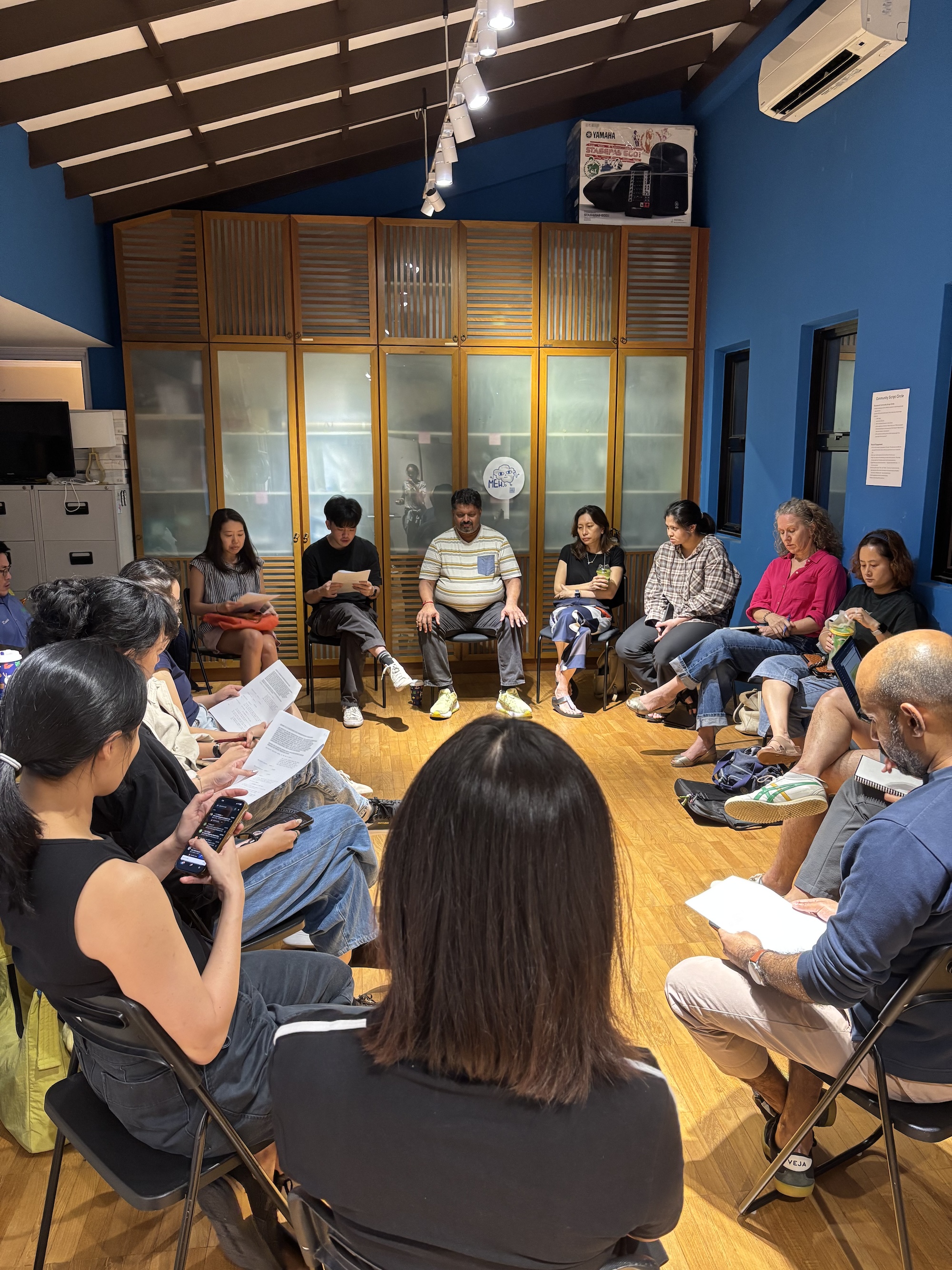 A large group of people seated in a circle. Some hold scripts in their hands.