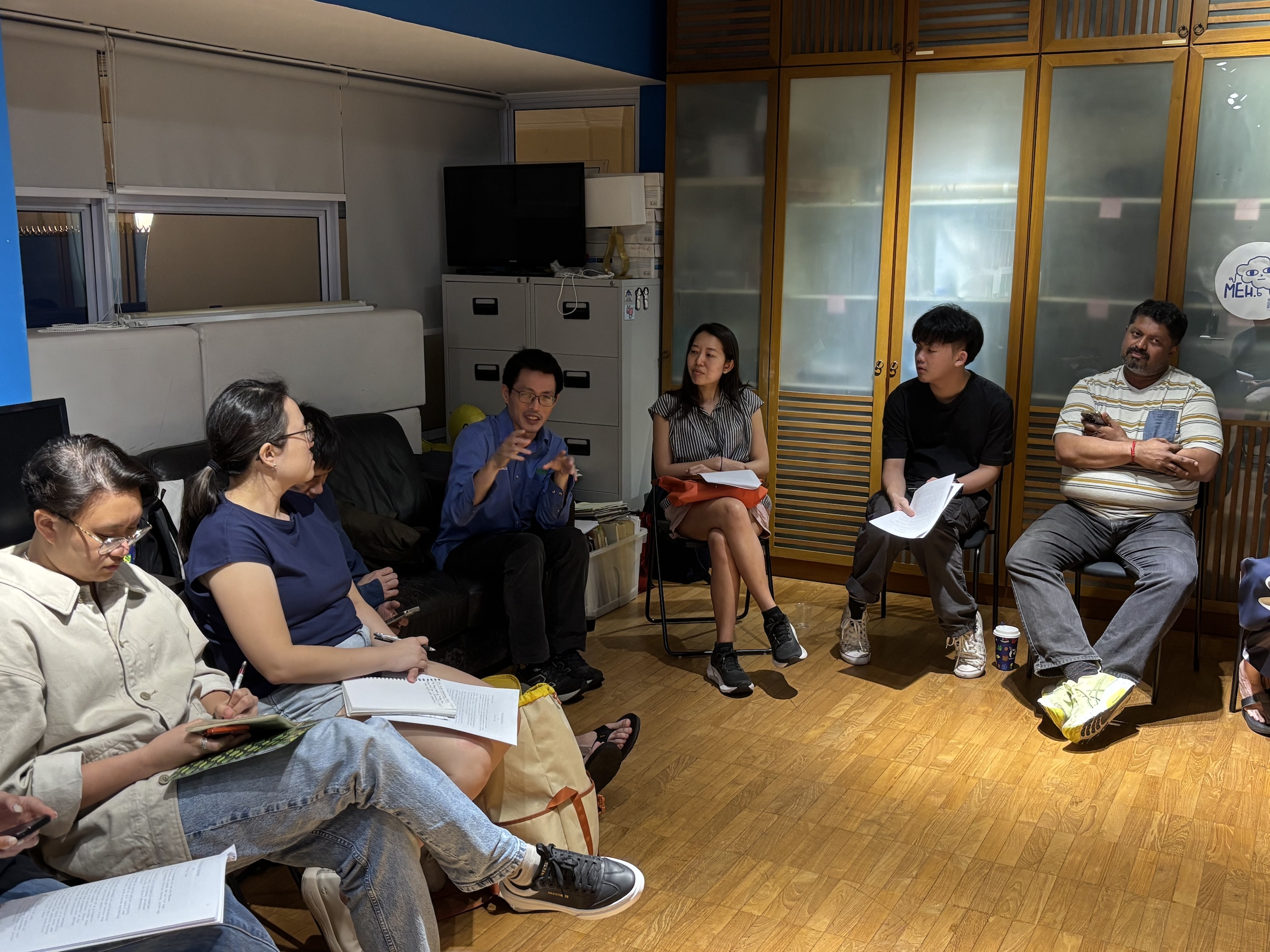 A male-presenting person seated in a circle, speaking and gesturing to others.