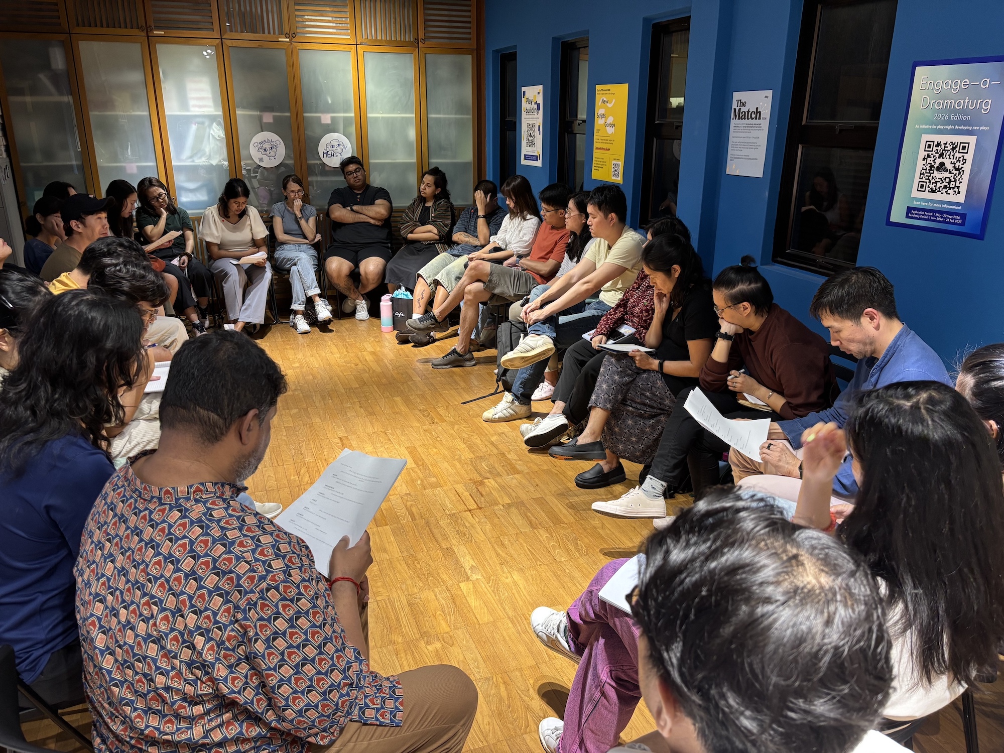 A large group of people seated in a circle. Some hold scripts.