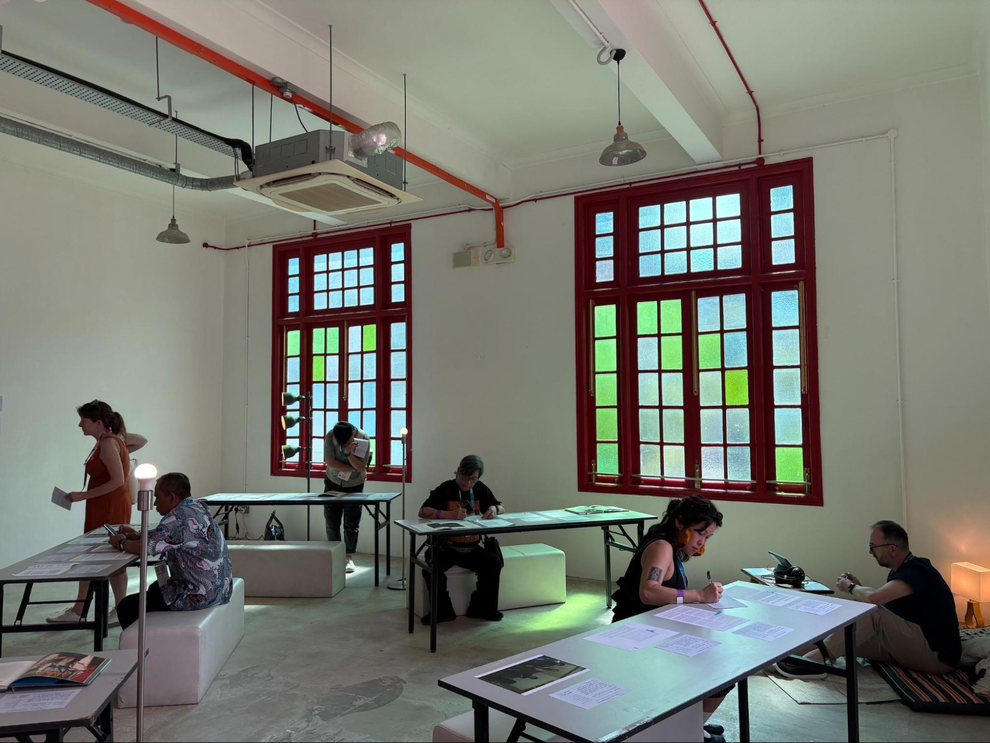 Six people scattered around a room with tables and benches, each writing on large slips of paper.