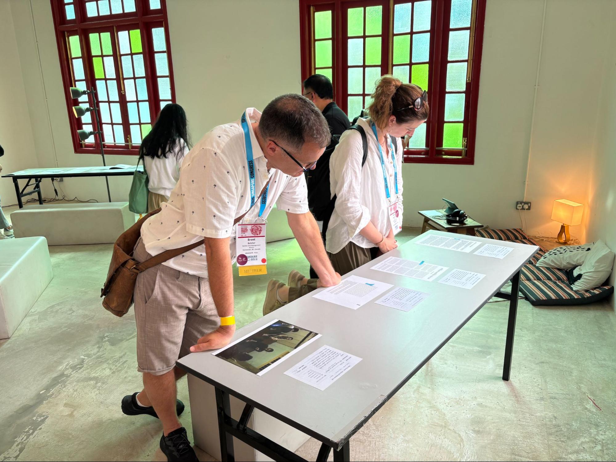 Two persons standing in front of a table, reading documents laid on its surface. Two persons standing in front of a table, reading documents laid on its surface.